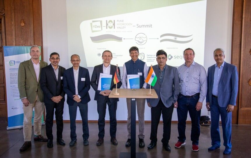 Deutsche und indische Delegationsmitglieder nach der Unterzeichnung am Stehpult vor der Summit-Projektion
