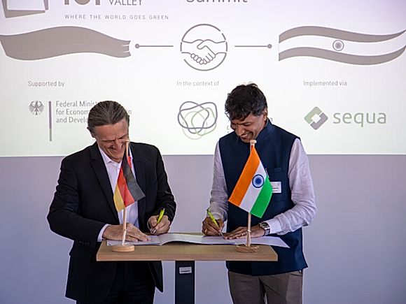 Zwei Vertreter unterzeichnen am Stehpult eine Vereinbarung, davor die deutsche und die indische Tischflagge