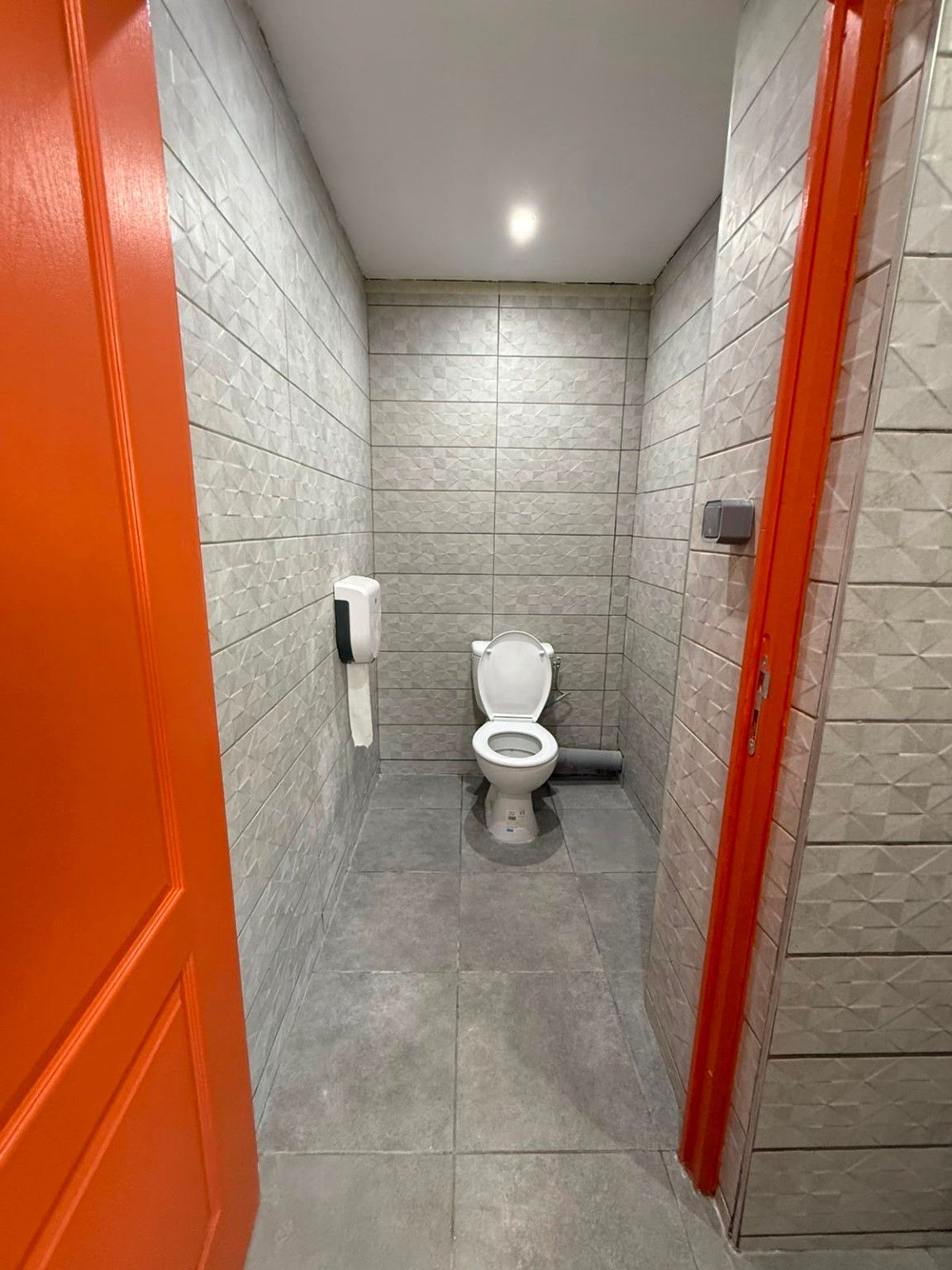 Cabine WC rénovée : faïence claire à relief, sol gris, WC neuf et porte orange