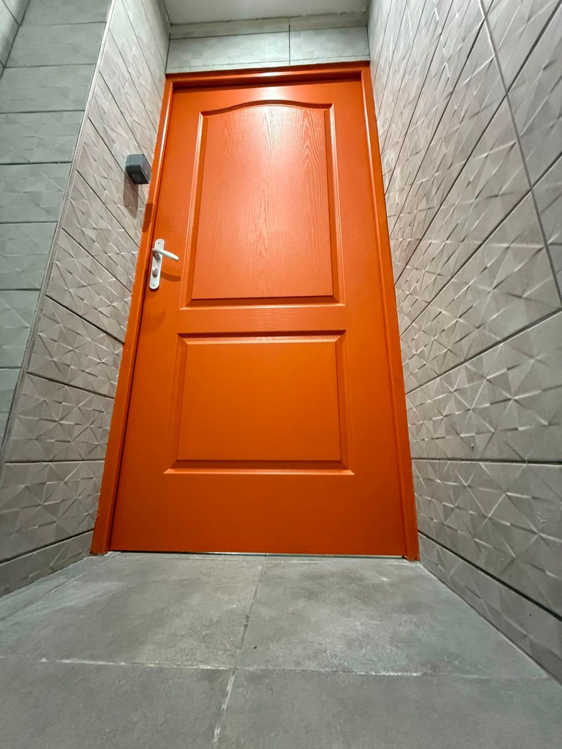 Porte de sanitaire repeinte en orange entre deux murs de faïence claire à relief