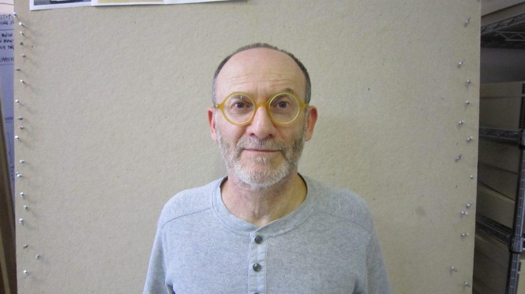 Danny Altman, in round yellow glasses and a gray henley, in front of a pinboard.