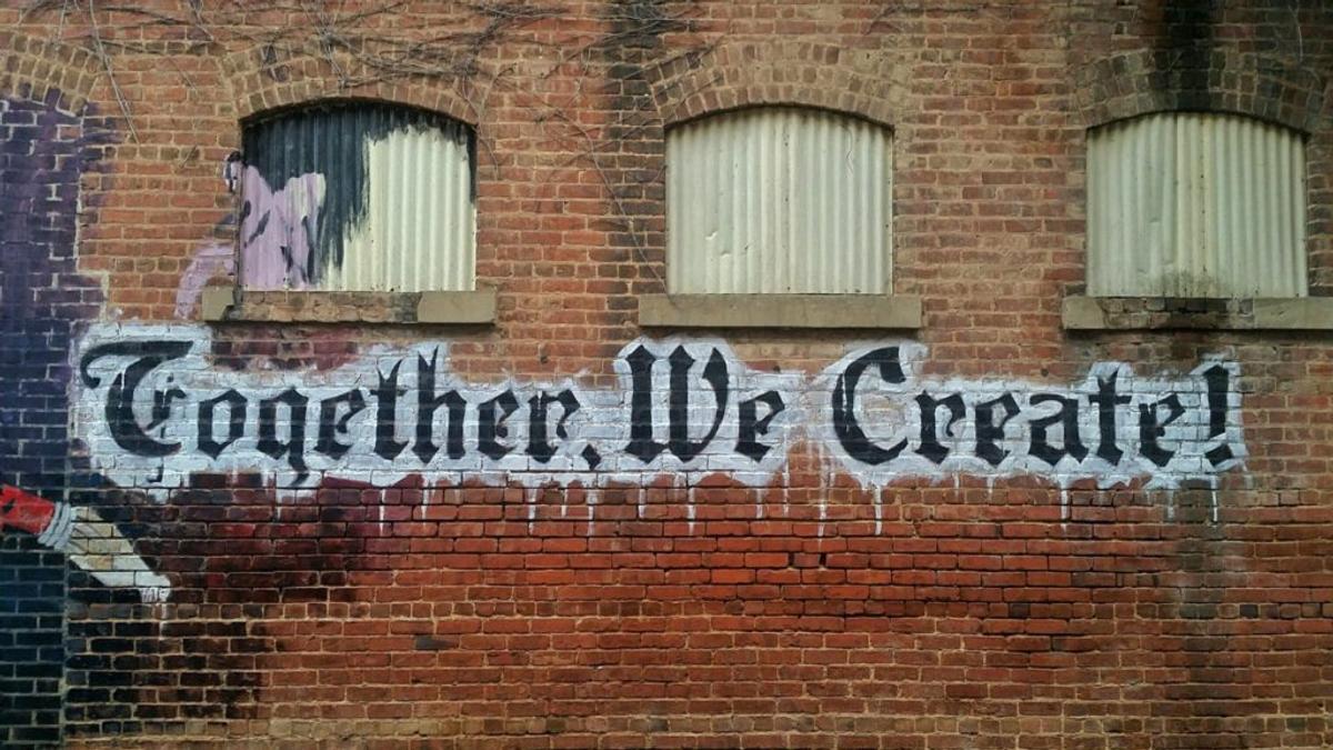 Blackletter graffiti on a brick wall reading "Together, We Create!"