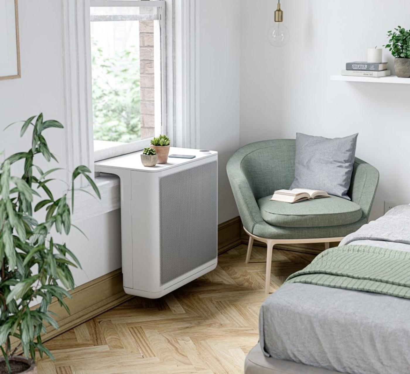 A white air purifier beside a bedroom window, armchair and bed alongside