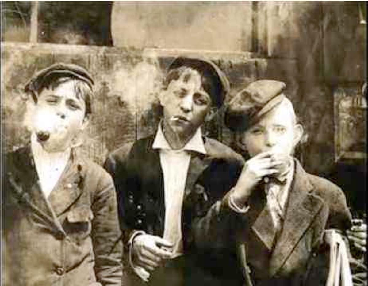 Three young newsboys in flat caps smoking cigarettes in an old sepia photograph.