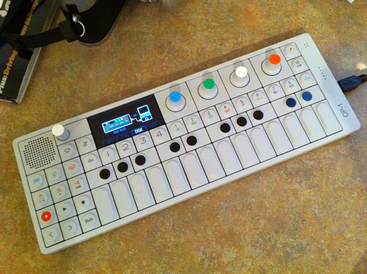 Teenage Engineering's OP-1 synthesizer, a slim white pocket synth with colored knobs and "OP-1" on its edge.
