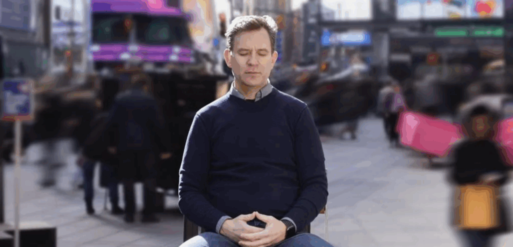 Dan Harris sitting with eyes closed and hands clasped, meditating in a blurred Times Square.