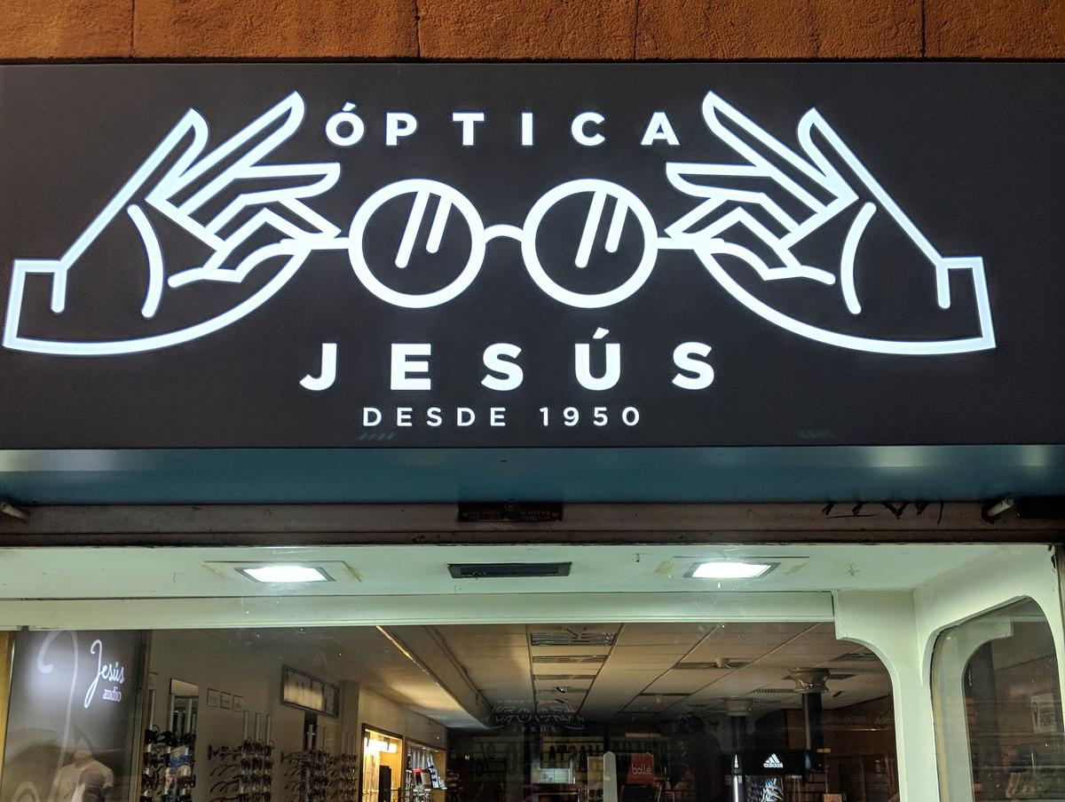 Backlit shop sign reading "Óptica Jesús, desde 1950," with two hands holding round spectacles.
