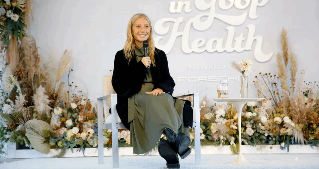 Gwyneth Paltrow onstage with a microphone before a wall reading "In Goop Health, presented by Porsche."