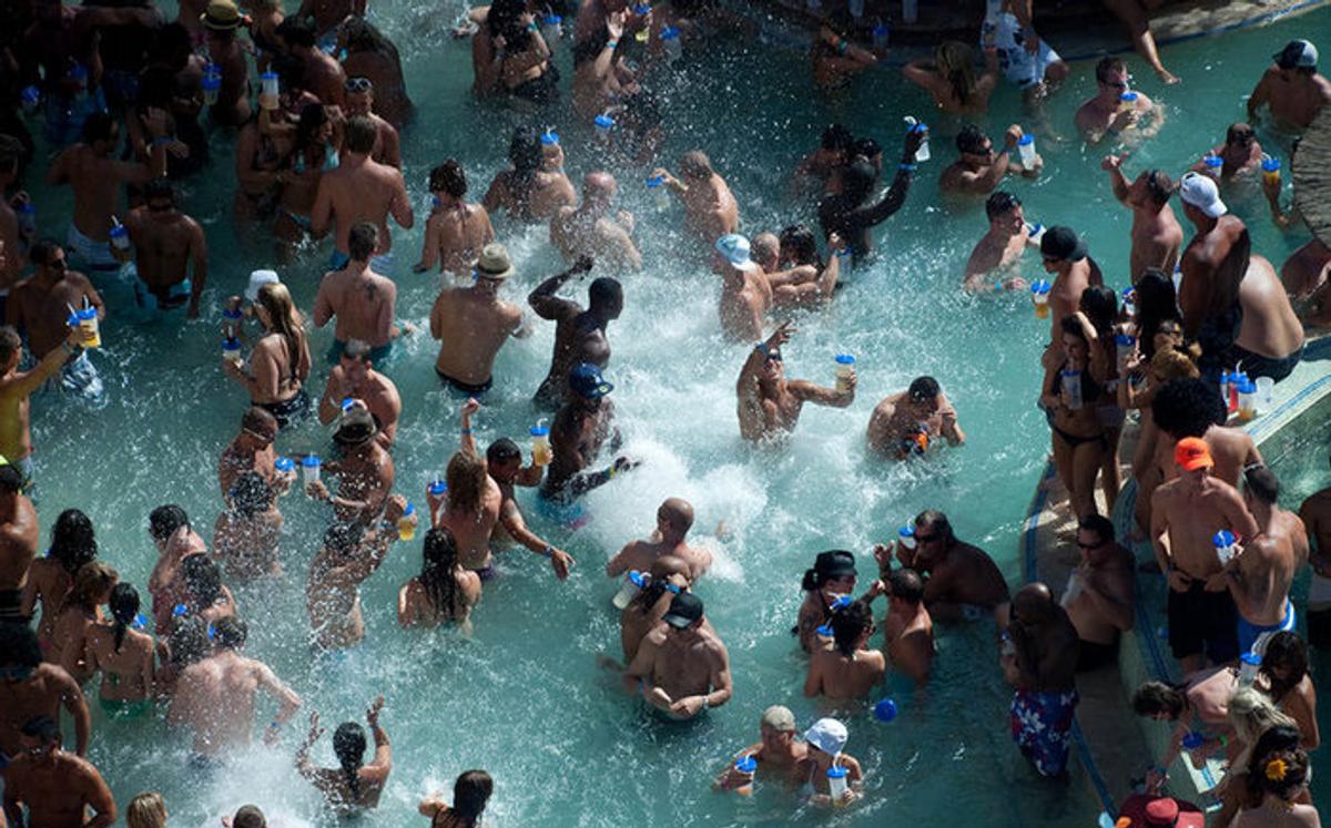 A packed hotel pool, dozens of people standing shoulder to shoulder in the water holding plastic cups.