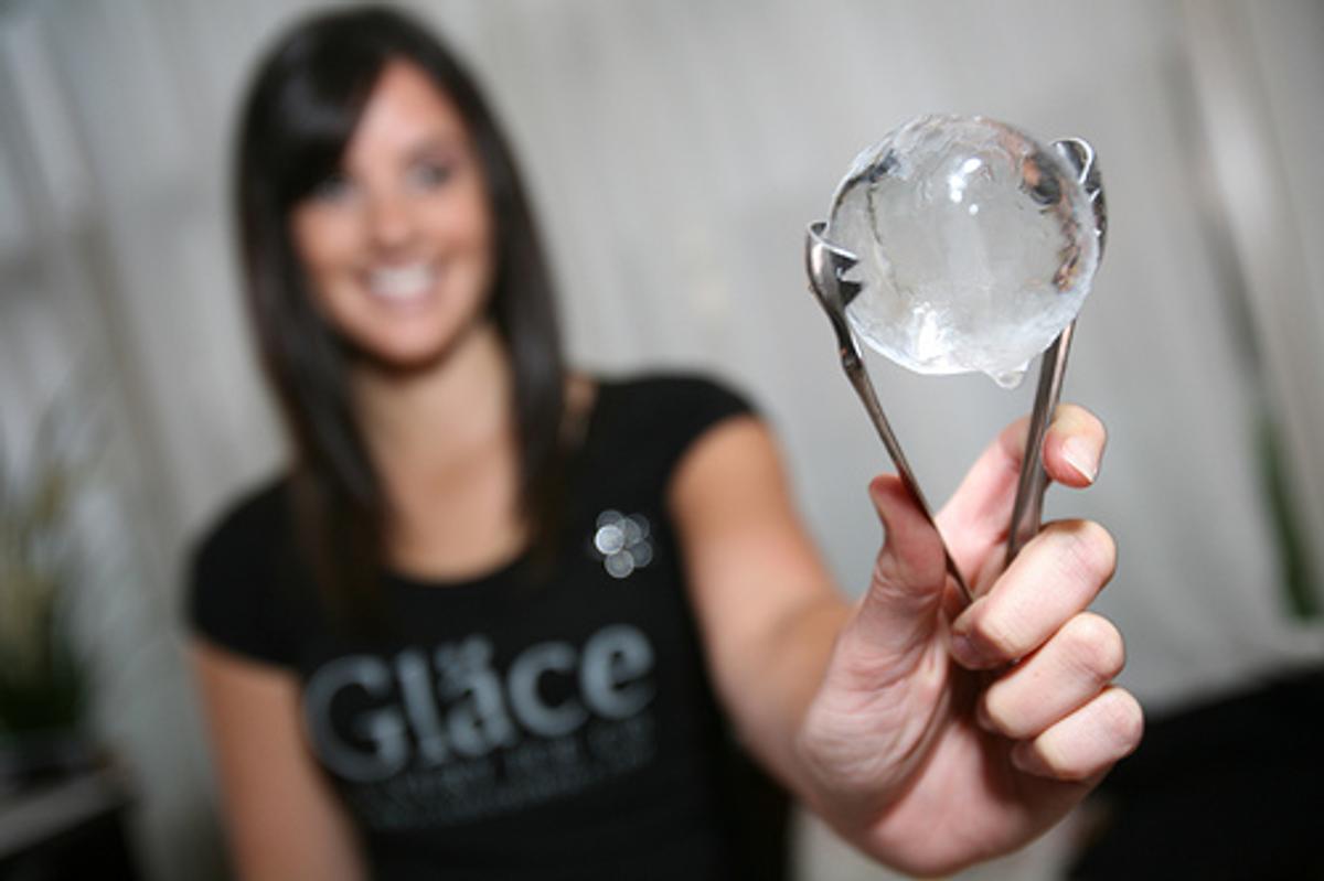 A woman in a "Gläce" T-shirt holds up a clear ice sphere with tongs.