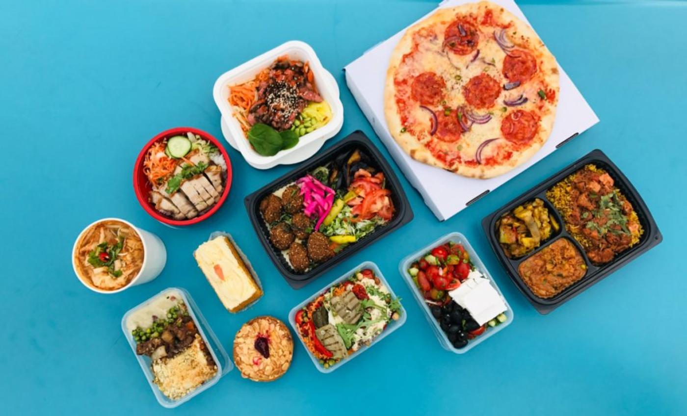 A spread of takeaway dishes on a bright blue surface — pizza, curries, salads, dumplings