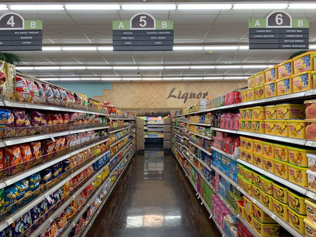 Supermarket snack aisle with overhead signs for aisle 5: "Snacks," "Chips," "Cookies / Crackers," "Candy," and "Liquor" painted on the back wall.