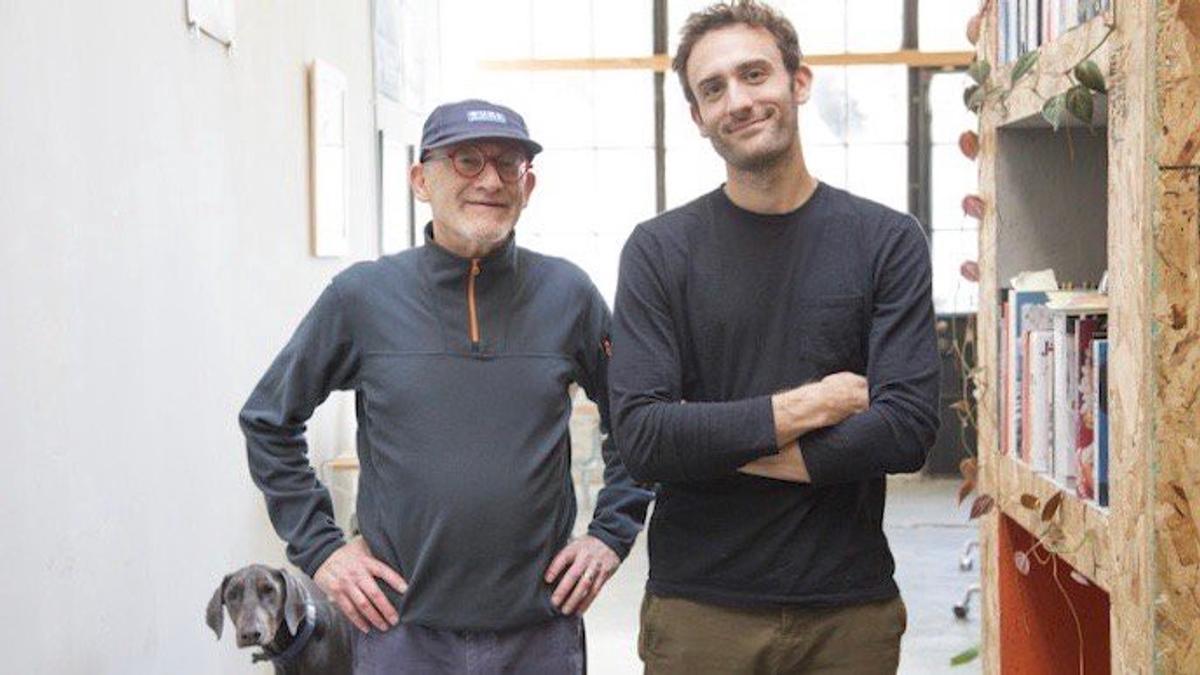 Two men standing side by side in a studio, an older man in a cap and glasses beside a younger bearded man with folded arms, with a dog at their feet.