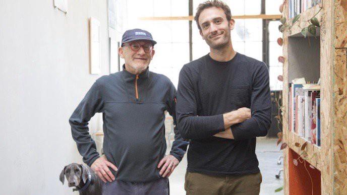 Two men standing side by side in a studio, an older man in a cap and glasses beside a younger bearded man with folded arms, with a dog at their feet.