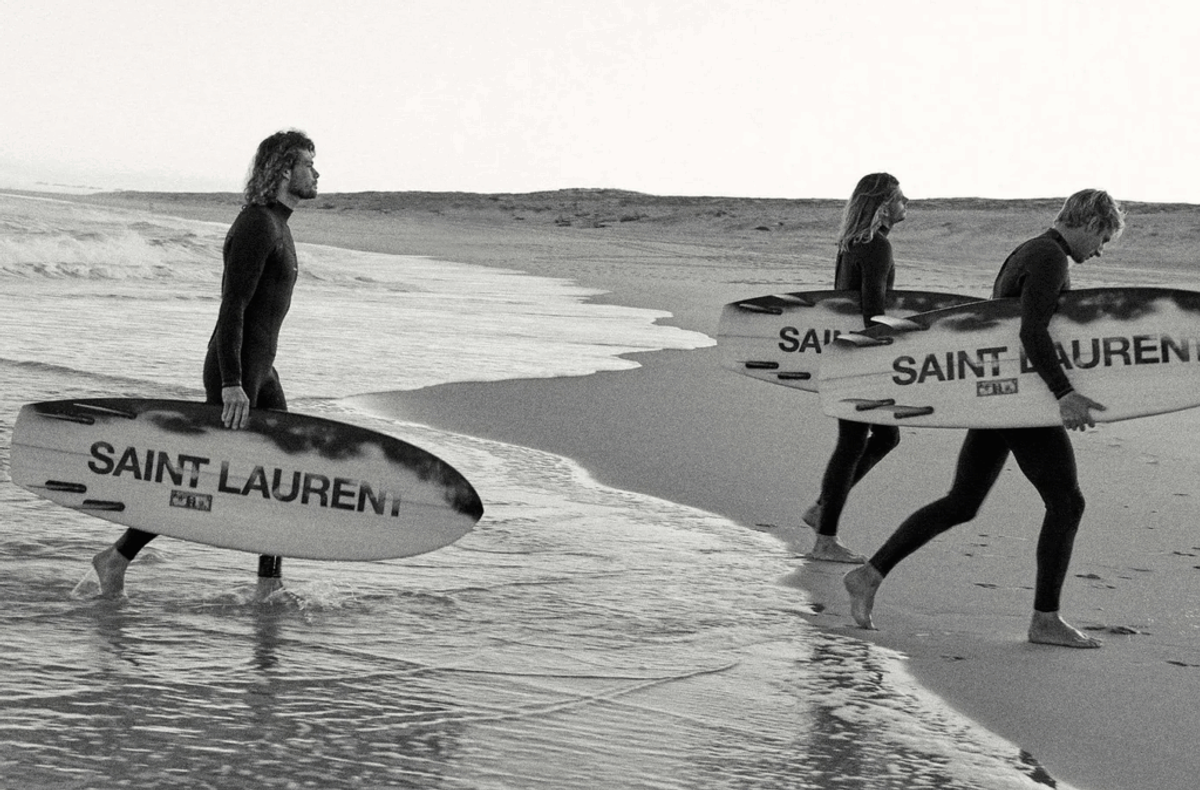 Three surfers in wetsuits carry boards branded "SAINT LAURENT" along a beach.