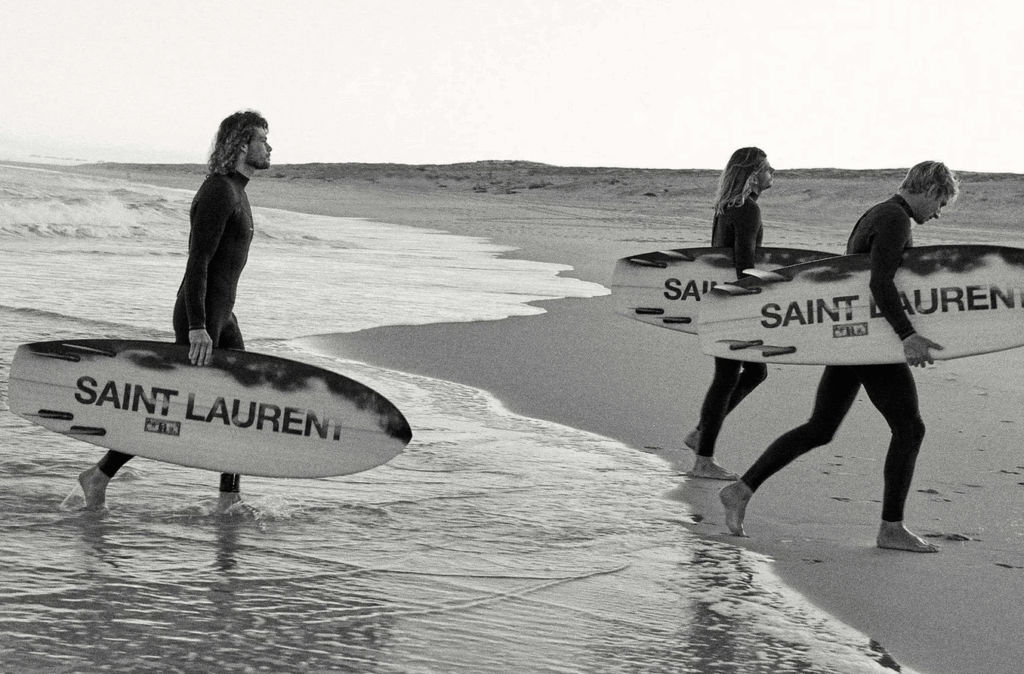 Three surfers in wetsuits carry boards branded "SAINT LAURENT" along a beach.