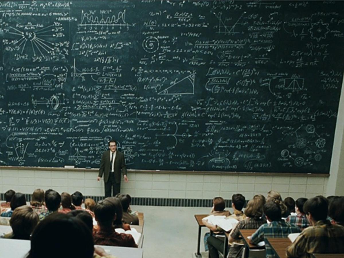 A lecturer stands before a huge blackboard covered in equations, facing a full class.