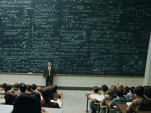 A lecturer stands before a huge blackboard covered in equations, facing a full class.