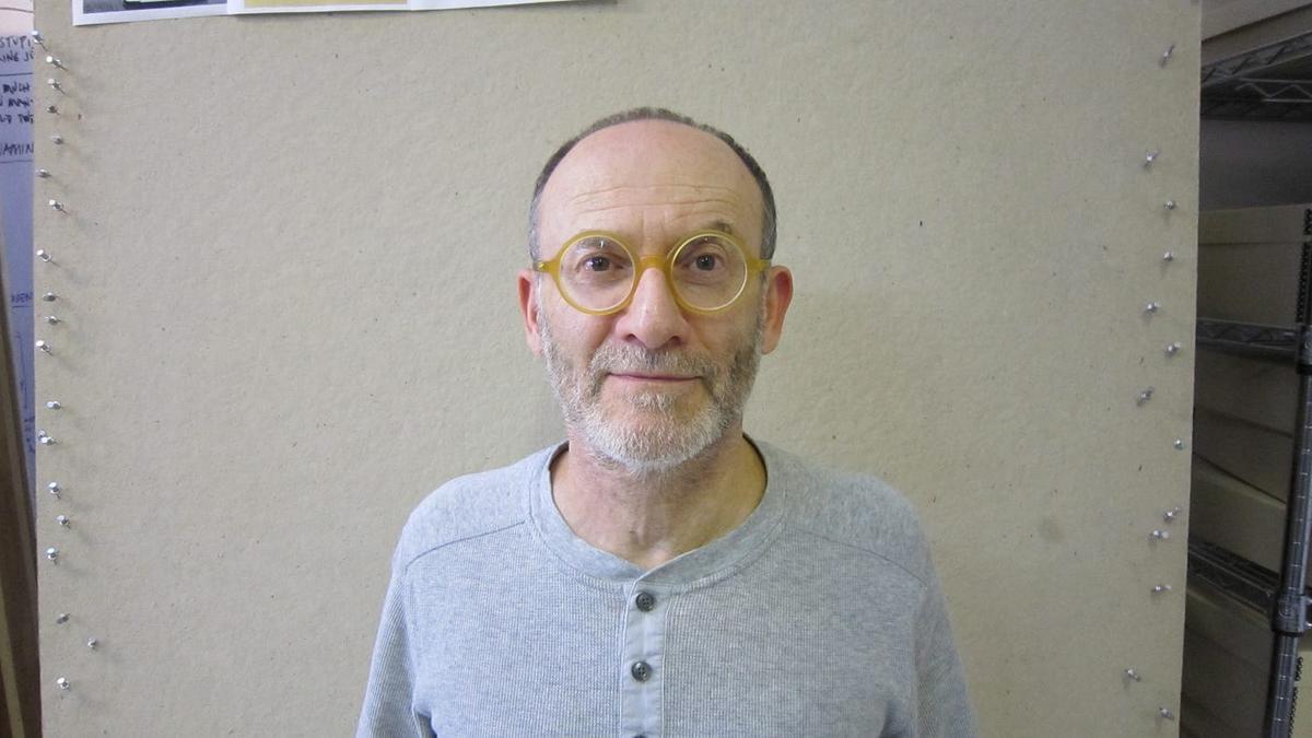 Danny Altman in round yellow glasses and a gray henley, standing against a corkboard wall.