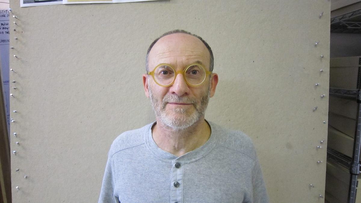 Danny Altman in round yellow glasses and a gray henley, standing against a corkboard wall.