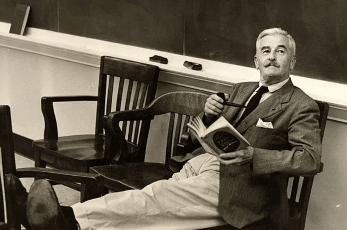 William Faulkner in a tweed jacket, feet up in a classroom chair, holding a pipe and an open book.