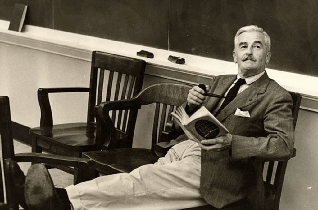 William Faulkner in a tweed jacket, feet up in a classroom chair, holding a pipe and an open book.