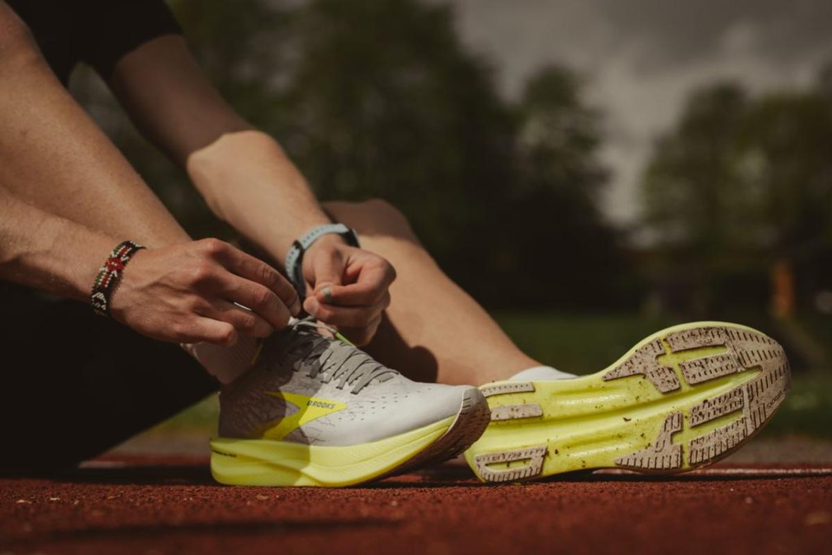 A runner laces up a gray and neon-yellow Brooks shoe on a red track.