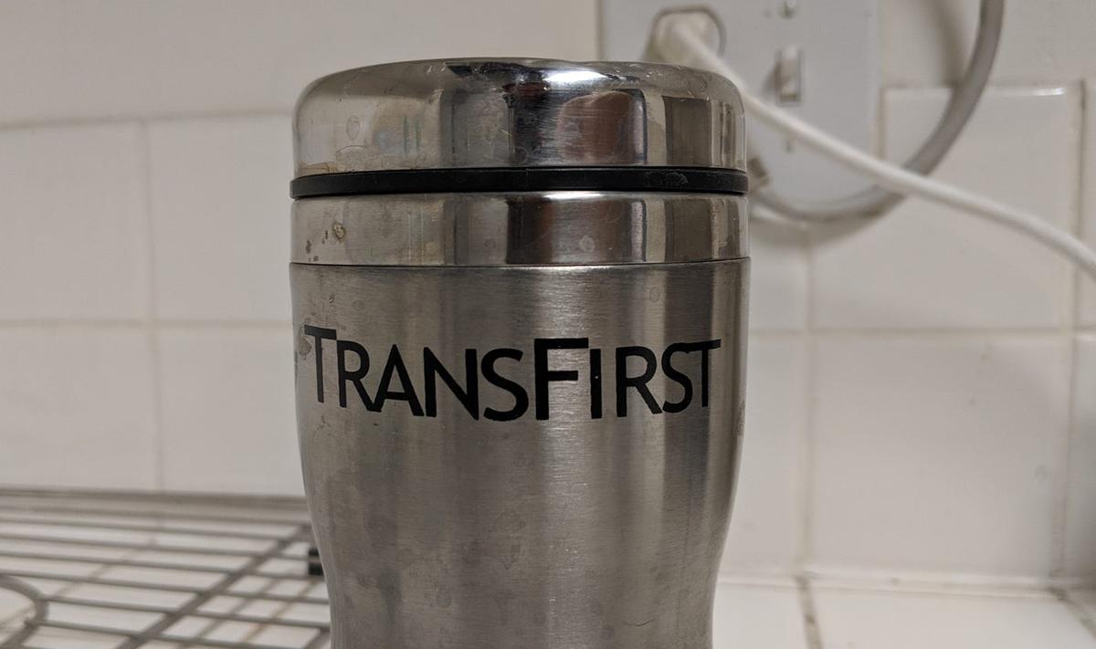 A scuffed stainless steel travel mug printed with the word "TransFirst".
