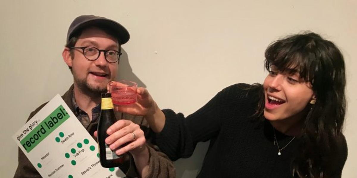 Two people toasting, one holding a scorecard reading "give the glory... record label!" listing Death Row, Sub Pop and Stones Throw.