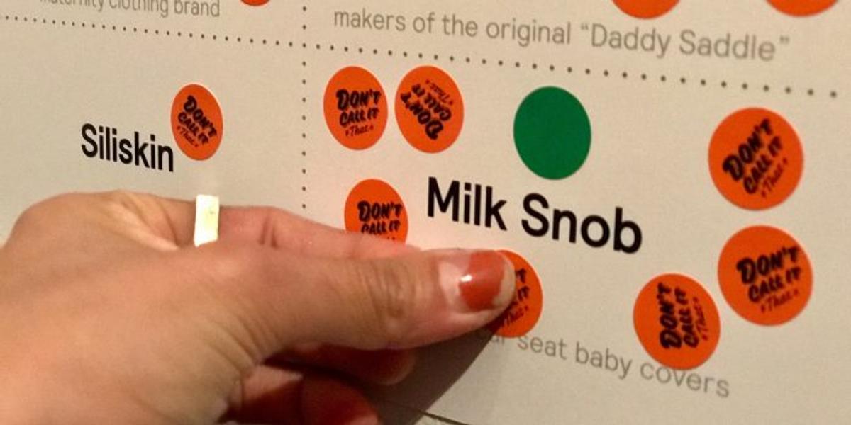 A hand placing orange "Don't call it that" stickers on a board listing baby brands including "Milk Snob," "Siliskin," and makers of the original "Daddy Saddle."