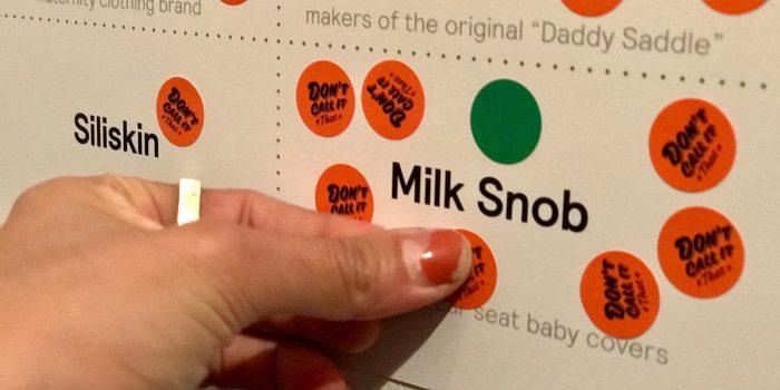 A hand placing orange "Don't call it that" stickers on a board listing baby brands including "Milk Snob," "Siliskin," and makers of the original "Daddy Saddle."