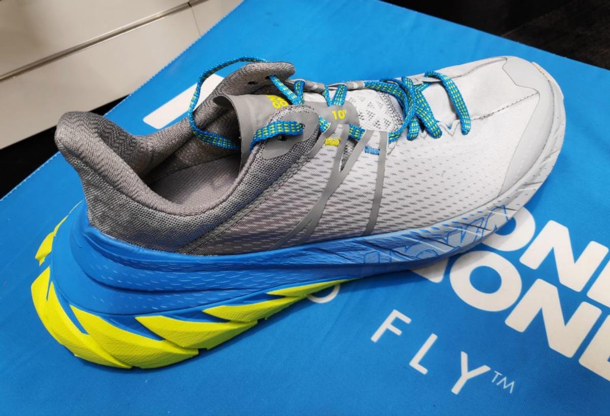A gray Hoka running shoe with thick blue and yellow cushioning, on a box reading "One One Fly."