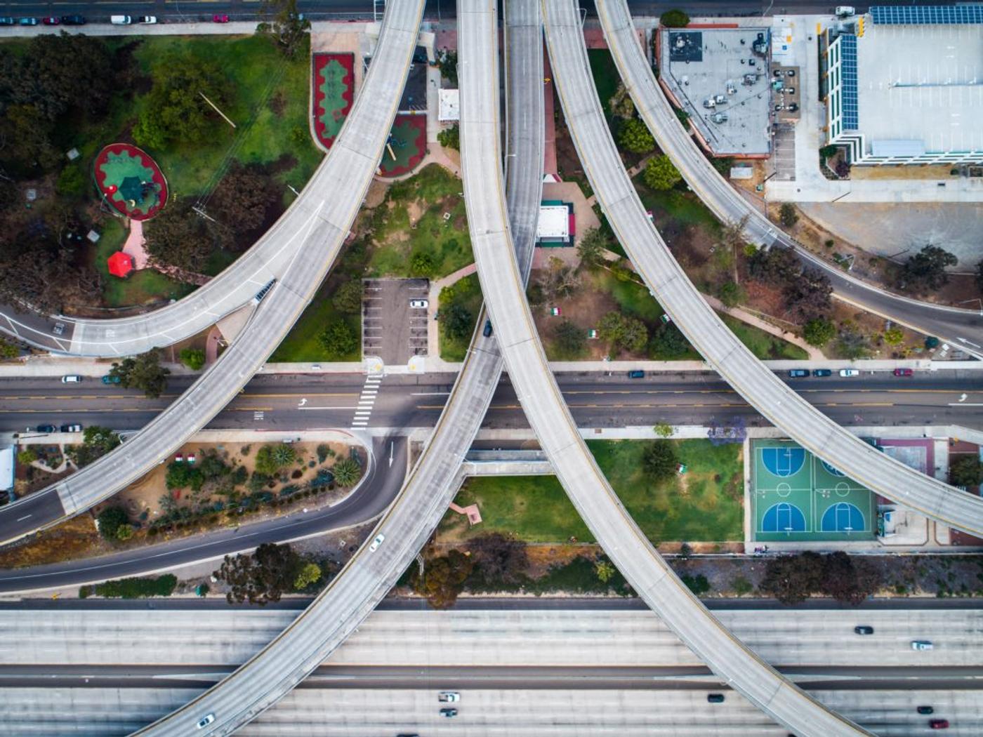 A freeway interchange from directly overhead, ramps curving past a park and courts