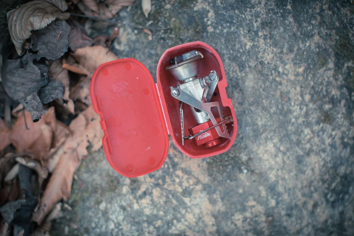 An MSR backpacking stove folded inside its small red plastic case, open on a rock.