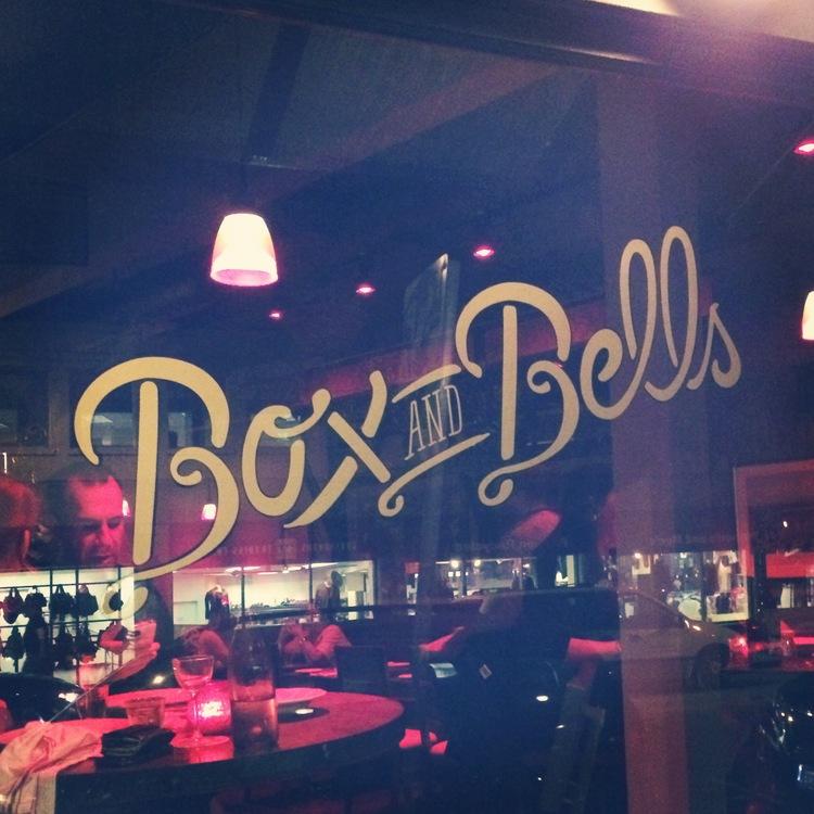 "Box and Bells" lettered in gold script on a restaurant window at night.