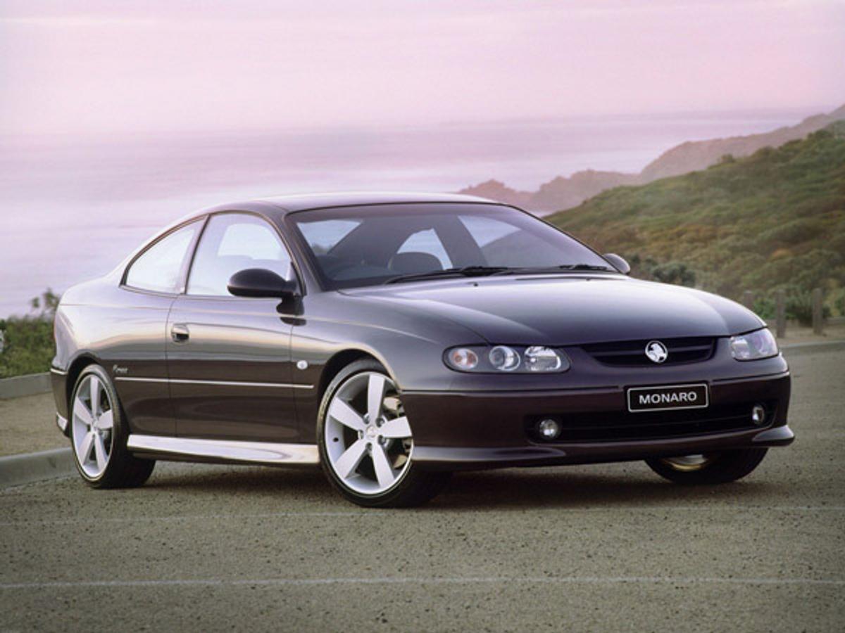 A dark purple Holden Monaro coupe parked on a coastal road, "MONARO" on its license plate.