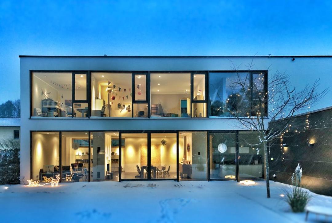A glass-fronted house lit from inside at dusk, snow across the garden