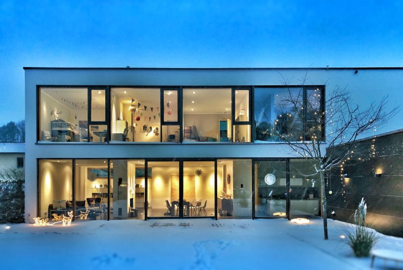 A glass-fronted house lit from inside at dusk, snow across the garden