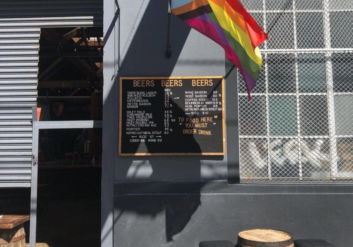 A brewery's hand-chalked beer board under a rainbow flag, barrels below