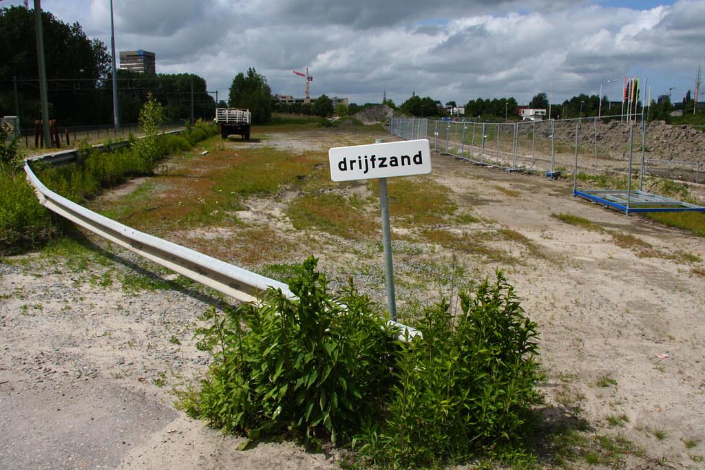 A white roadside sign reading "drijfzand" — Dutch for quicksand — on a weedy vacant lot.