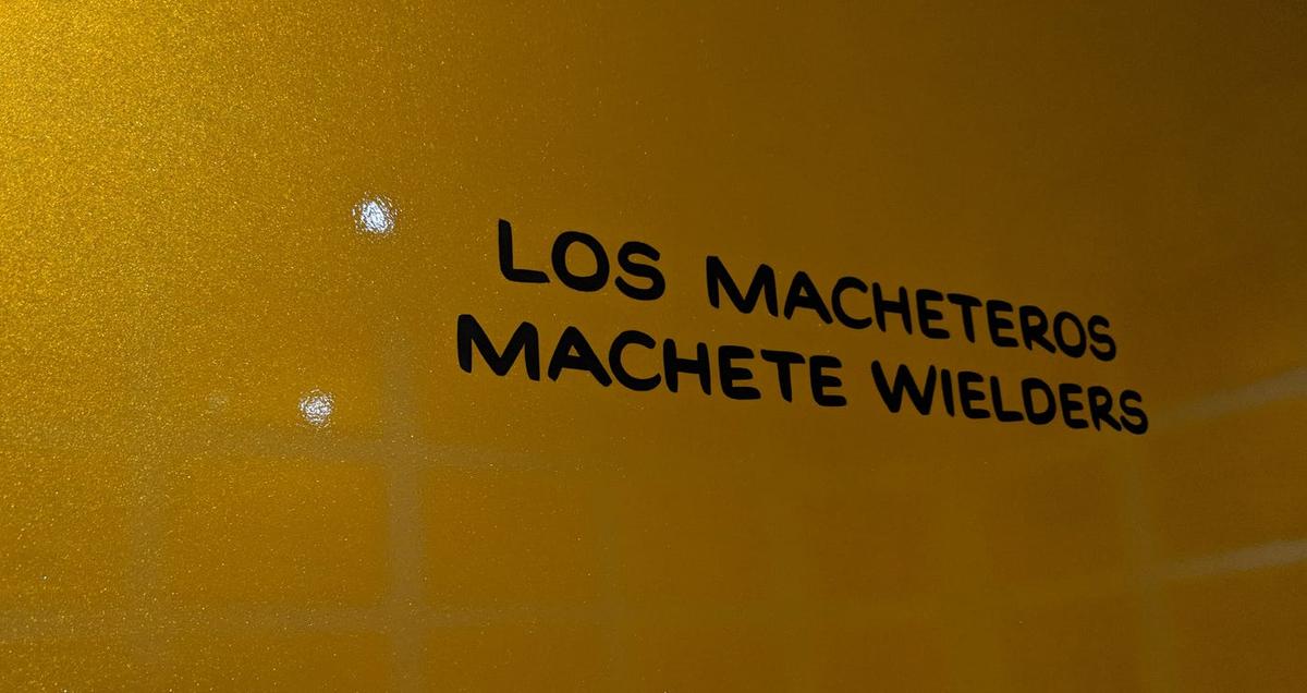 Black lettering on a gilded panel reading "Los Macheteros / Machete Wielders."