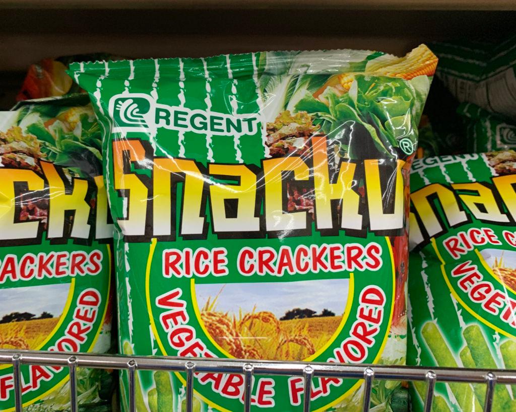 Green bags of Regent "Snacku" rice crackers, vegetable flavored, on a store shelf.