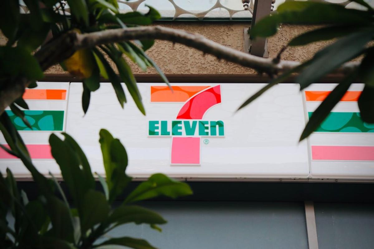 A 7-Eleven storefront sign seen through tree leaves.