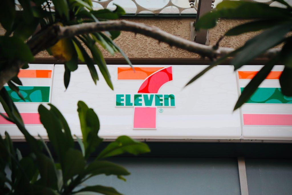 A 7-Eleven storefront sign seen through tree leaves.