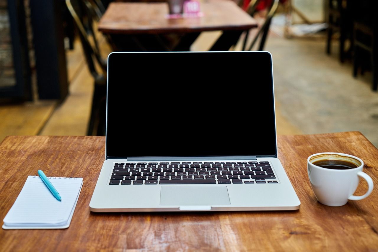 An open laptop on a wooden cafe table next to a coffee cup and a notepad with a pen.