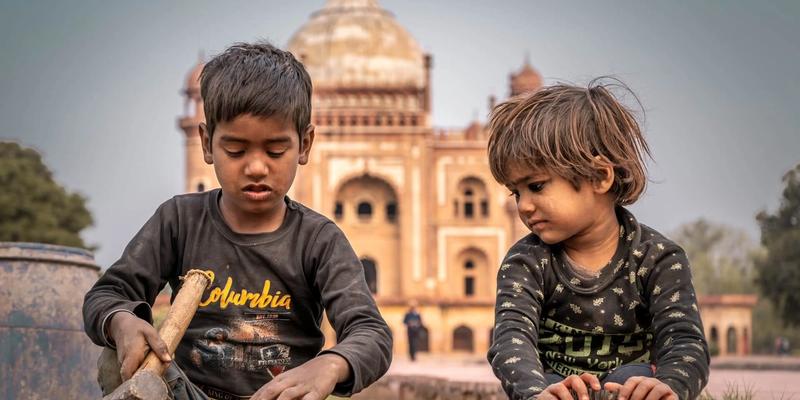 Two young children sitting on the ground in Delhi, with Safdarjung's Tomb behind them.