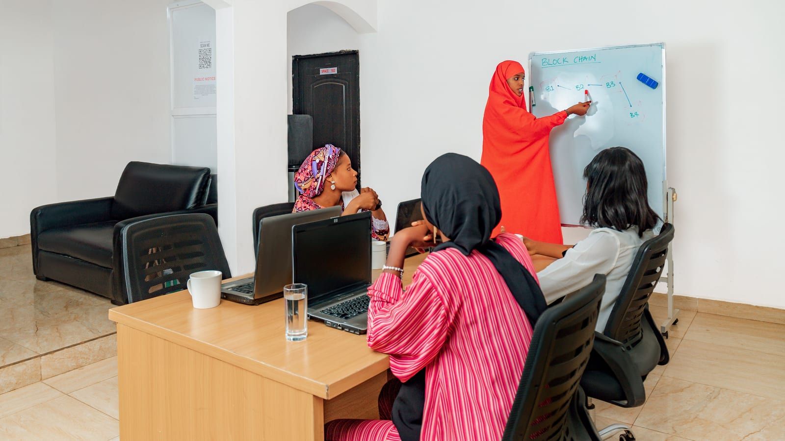 Women working at laptops while a trainer writes on a whiteboard.