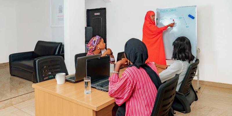 Women working at laptops while a trainer writes on a whiteboard.