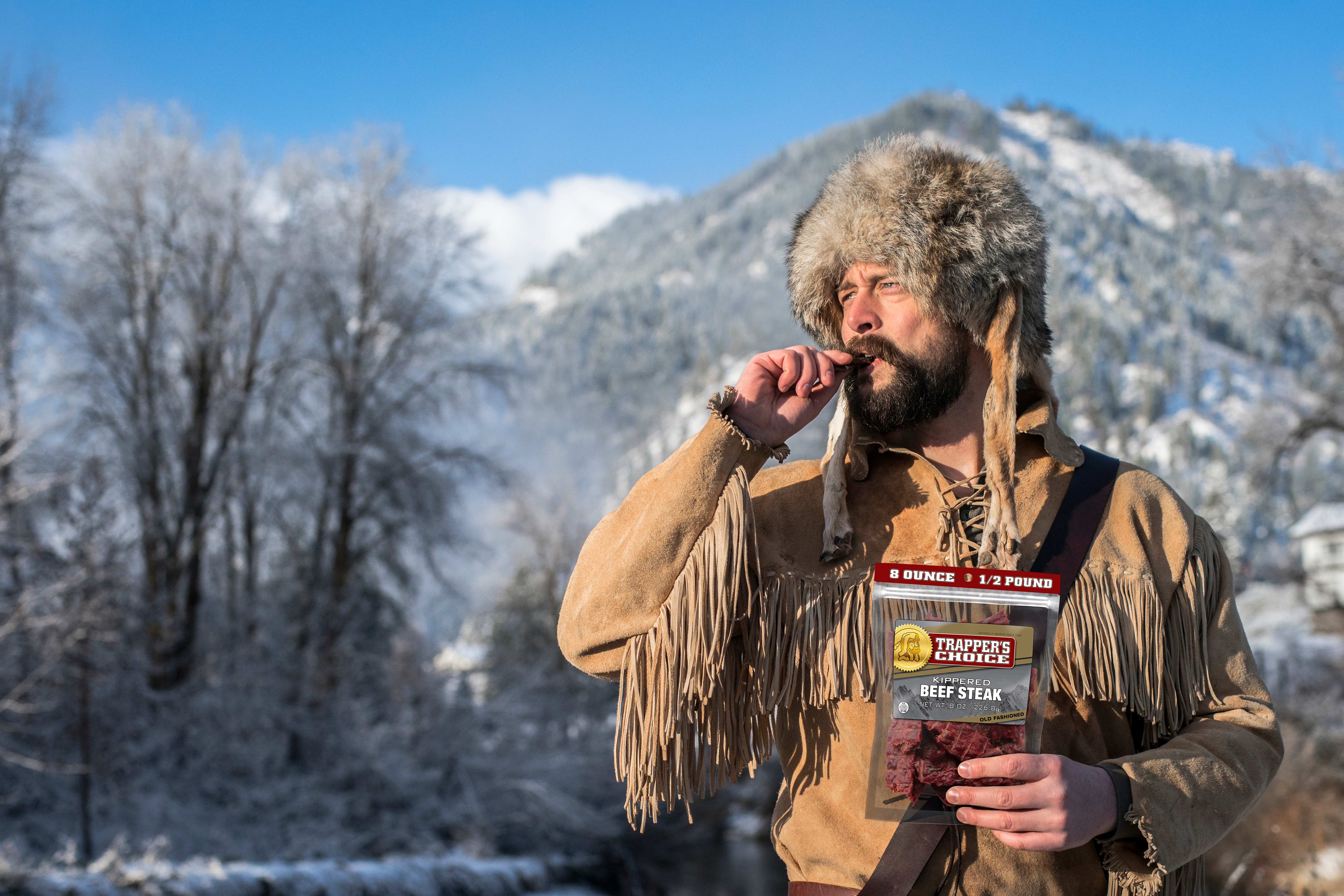 old trapper eating beef steak outside with a mountain behind him