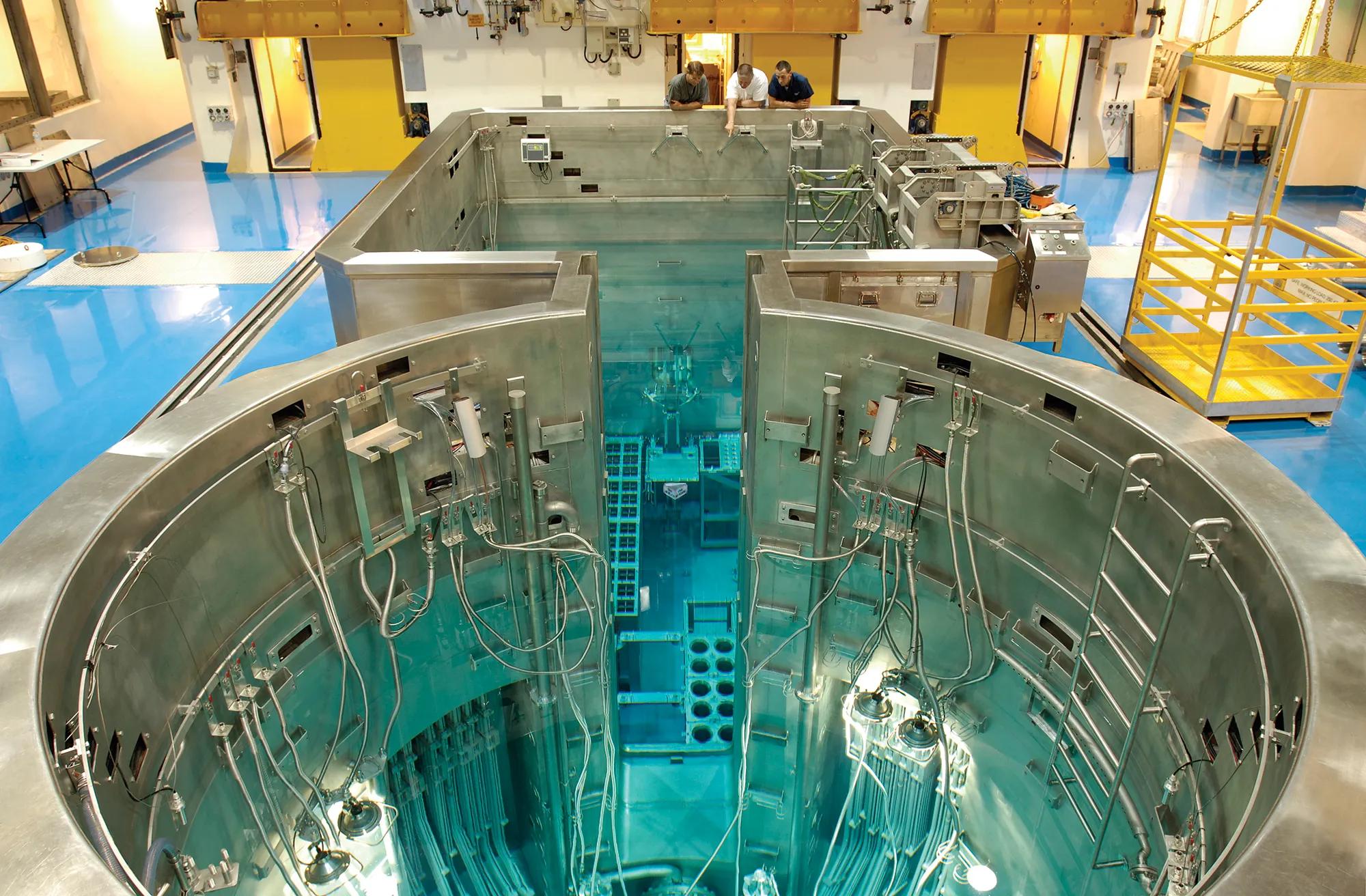 Interior of a nuclear research reactor facility showing a large stainless-steel reactor pool filled with clear blue water, with submerged reactor components, cables, and instrumentation visible, and technicians observing from a platform above.